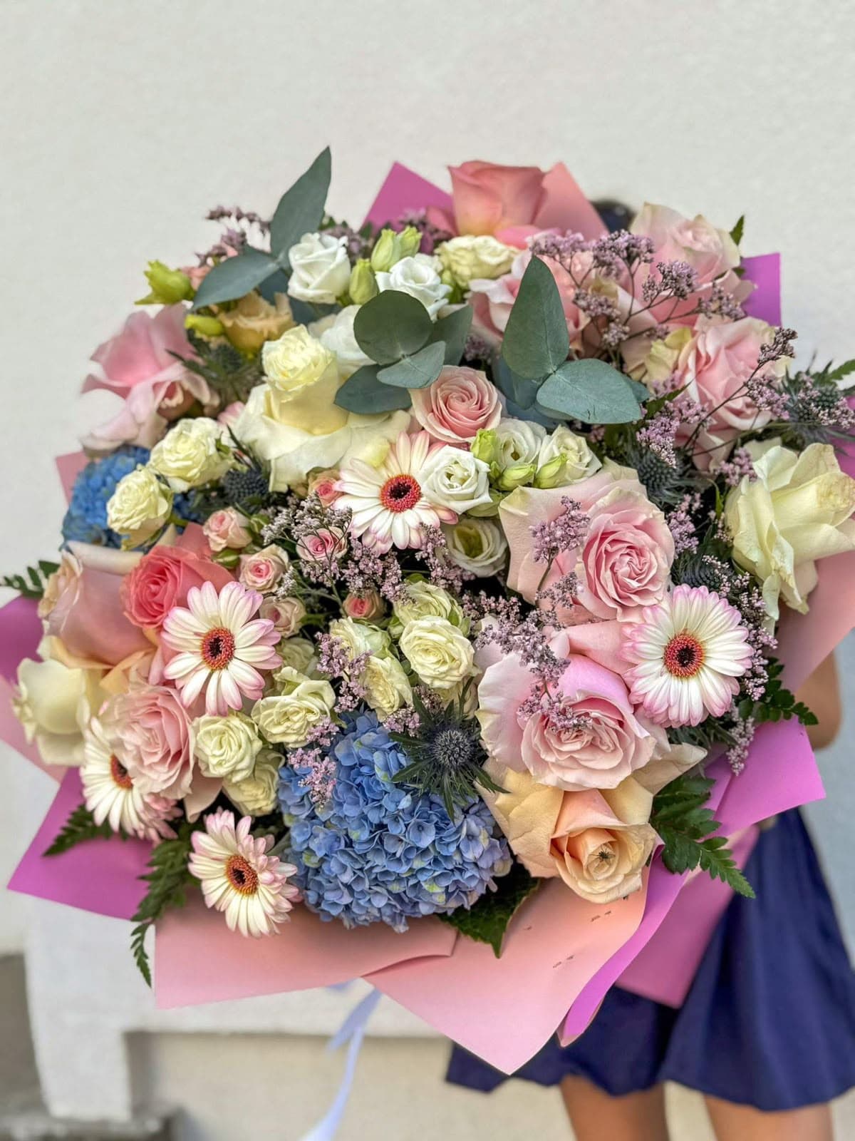 Buchet elegant cu trandafiri roz și crem, hortensii albastre, gerbera roz pal și flori delicate de sezon, învelit în hârtie decorativă roz fucsia. Aranjament floral premium, ideal pentru aniversări, ocazii speciale și cadouri elegante.