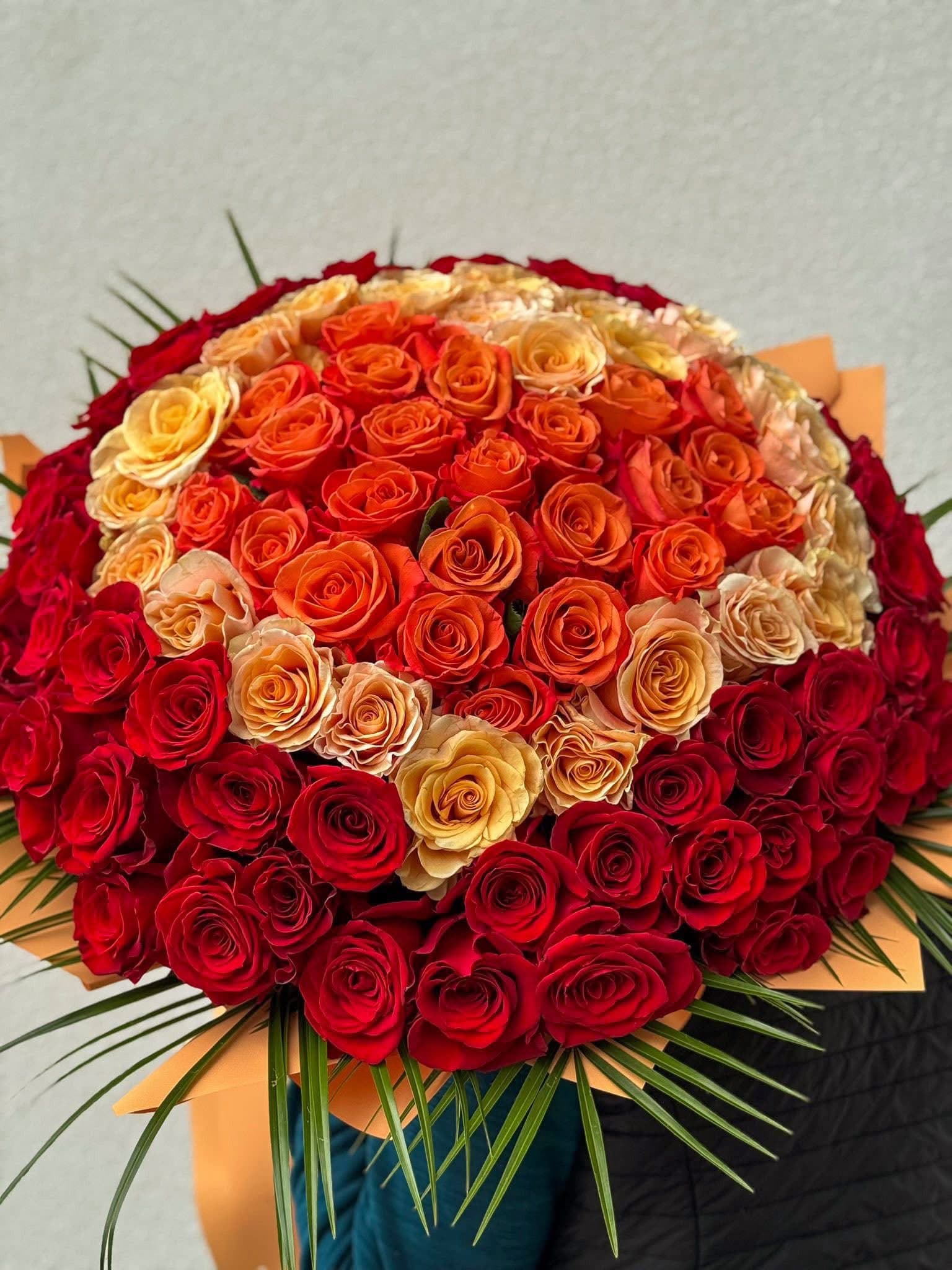 Buchet spectaculos din trandafiri roșii, portocalii și galbeni, aranjați în degrade cromatic și completați cu frunziș verde decorativ. Un buchet floral premium, ideal pentru declarații de dragoste, aniversări și ocazii memorabile.