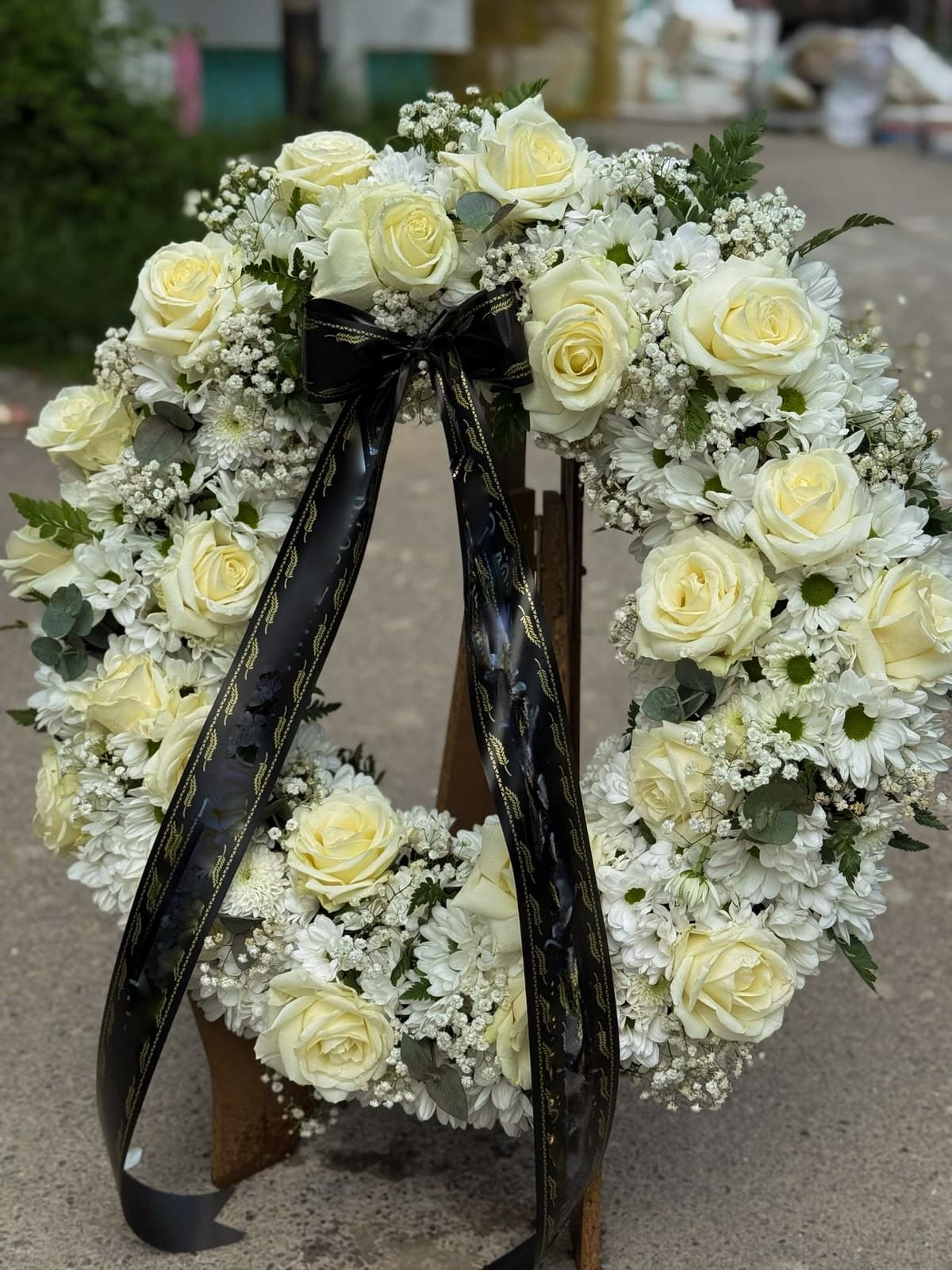 Coroană funerară elegantă din trandafiri ivoire, crizanteme albe, gypsophila și eucalipt, decorată cu o panglică neagră cu detalii aurii. Un omagiu floral delicat și solemn, potrivit pentru a transmite respect, compasiune și un ultim rămas-bun.