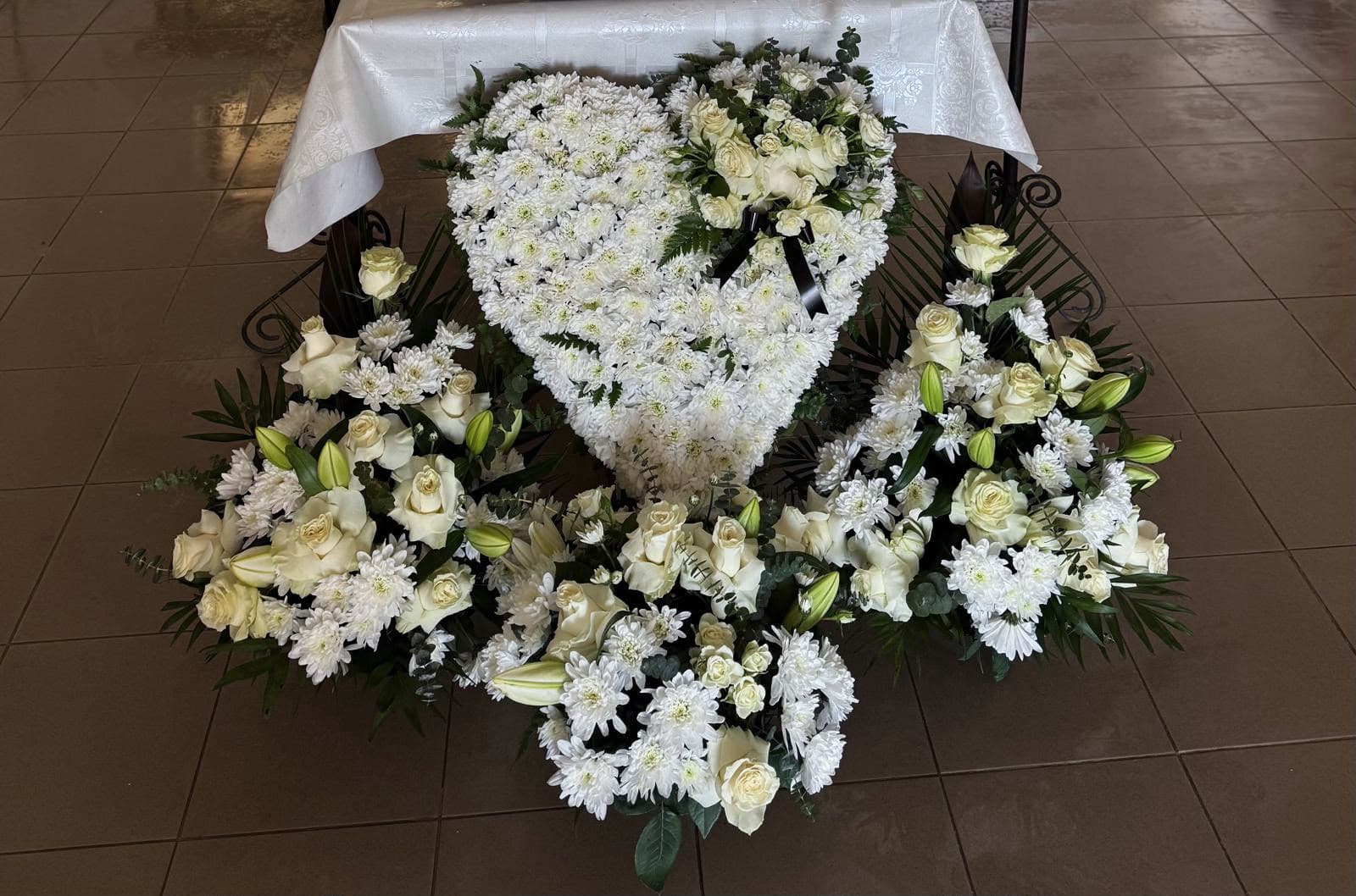 Aranjament funerar elegant în nuanțe de alb și ivoire, cu trandafiri, crizanteme și crini, completat de frunziș verde și accente decorative. Un omagiu floral solemn, potrivit pentru ceremonia de rămas-bun și pentru a exprima respectul și compasiunea față de cei dragi.