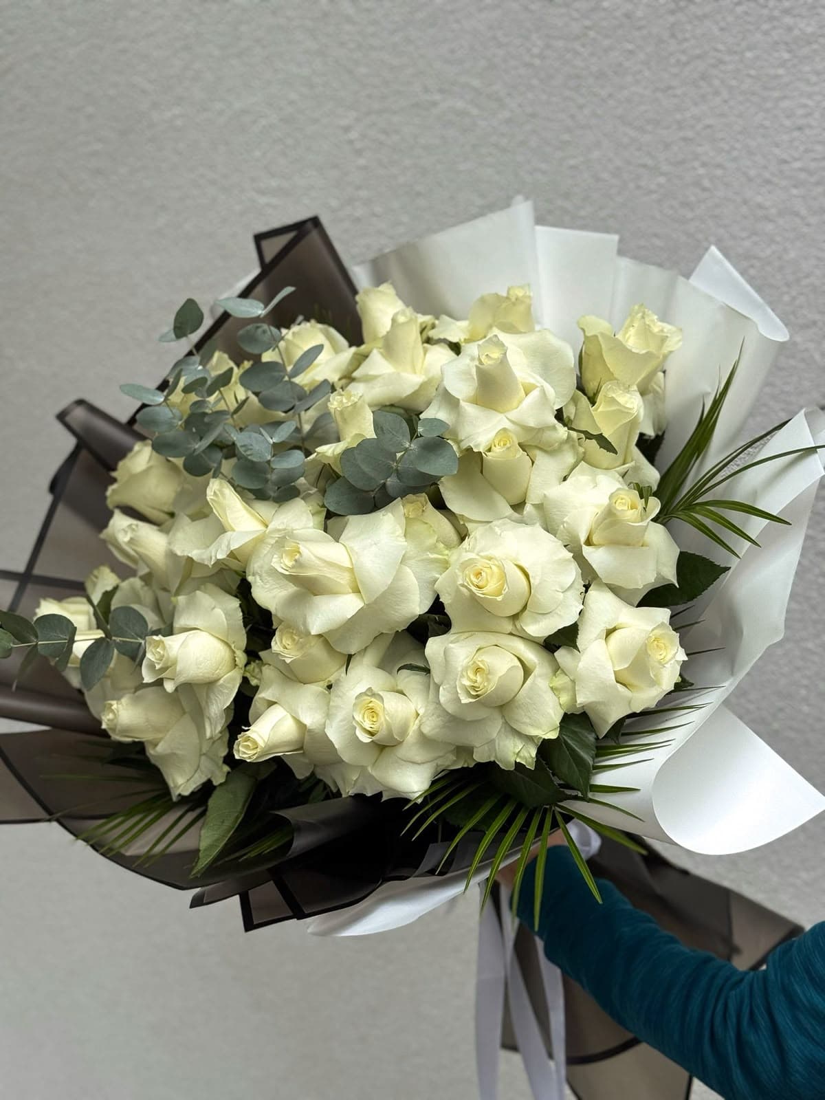 Buchet elegant din trandafiri albi ivoire, completat cu eucalipt argintiu și frunziș decorativ, învelit în hârtie albă și neagră. Un aranjament floral rafinat, potrivit pentru ceremonii funerare, omagii de rămas-bun și exprimarea respectului față de cei dragi.